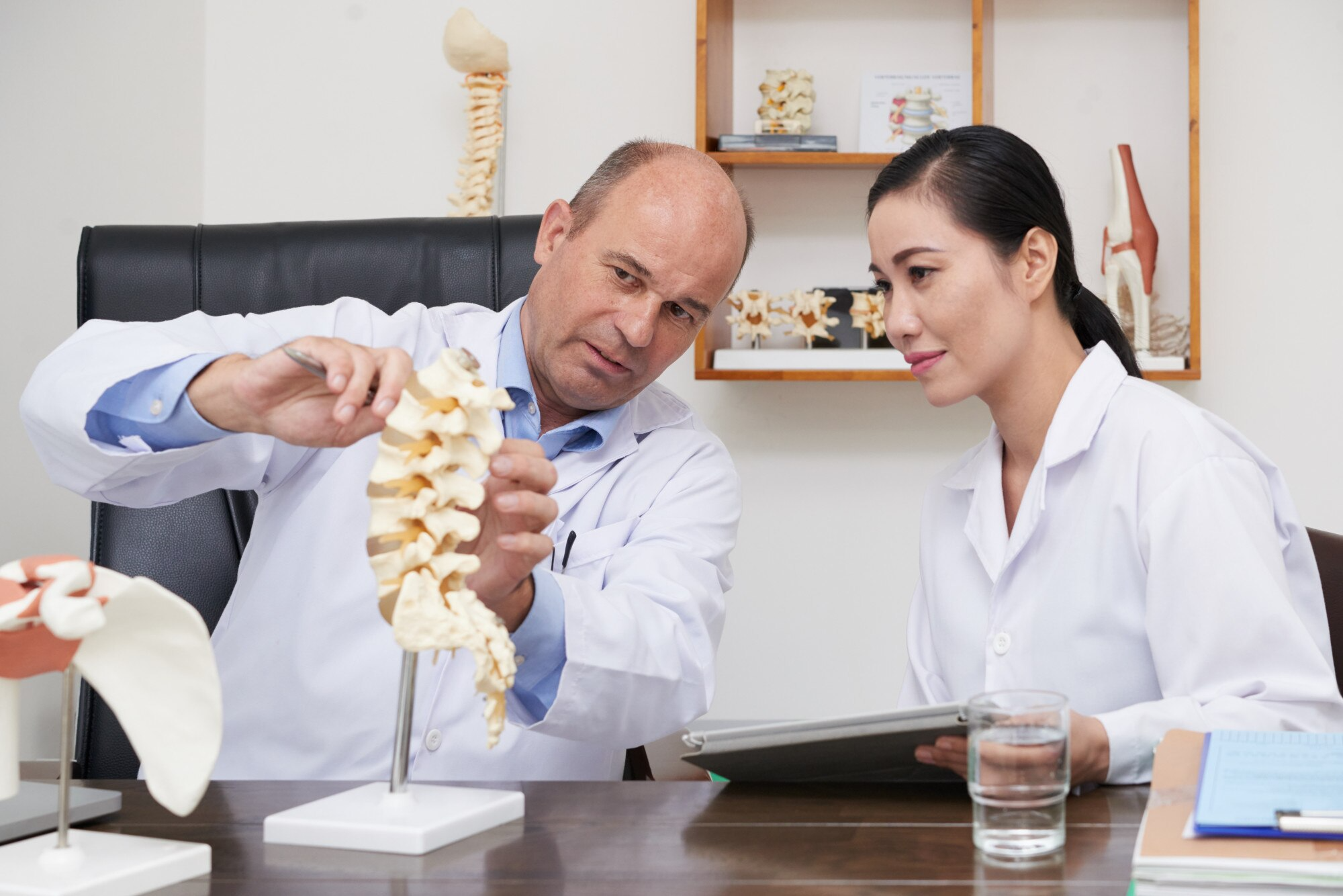 Two doctors and a chiropractor examine a spine model at a desk, with medical books and anatomical models on shelves in the background, discussing possible treatments for back pain following a car accident.