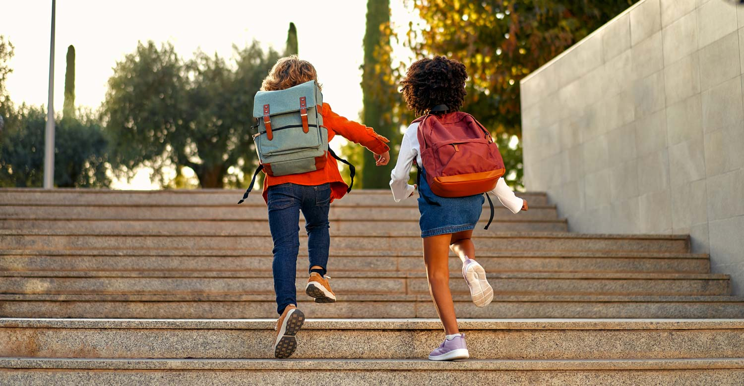 Two children with backpacks run up a flight of outdoor stairs surrounded by trees, completely carefree and oblivious to the world's troubles, like back pain or a recent car accident.