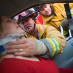Two firefighters assist an injured woman wearing a neck brace inside a vehicle, likely from a car accident. One firefighter supports her head to prevent further injury, while the other observes and prepares equipment for any potential back pain issues.