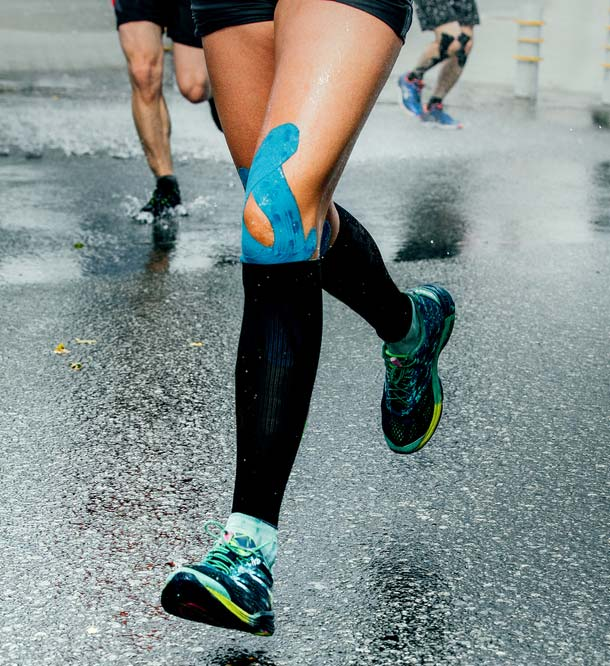 Close-up of a runner's legs on a wet pavement with blue kinesiology tape on the left knee. Other runners and splashing water visible in the background, reminiscent of someone pushing through back pain after consultation with a chiropractor.
