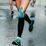Close-up of runners’ legs during a race on a wet road. One runner’s leg has blue kinesiology tape applied, perhaps a precaution by their chiropractor after recovering from back pain. They are wearing black compression sleeves and blue-green running shoes.