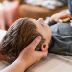 A person gently cradles a child's head while the child lies down on a bed, seeking relief from back pain after a minor car accident.