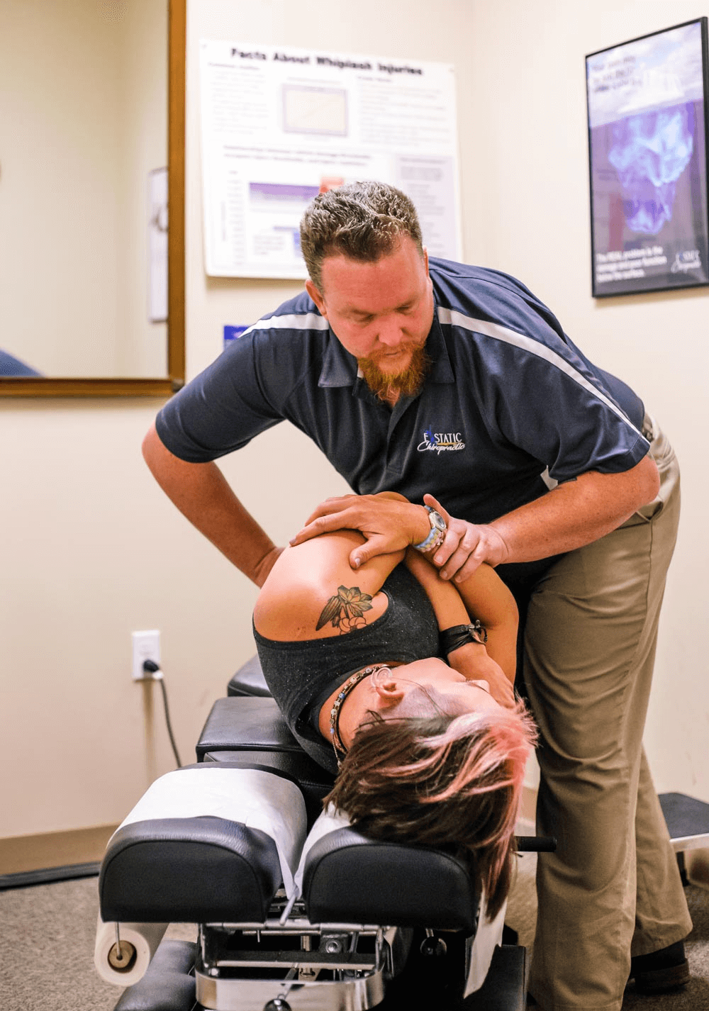 A chiropractor adjusts a patient lying on their side on an adjustment table. The patient, who is seeking relief from back pain following a car accident, has a tattoo on their upper arm and is wearing a sleeveless top. Chiropractic posters are visible on the wall.