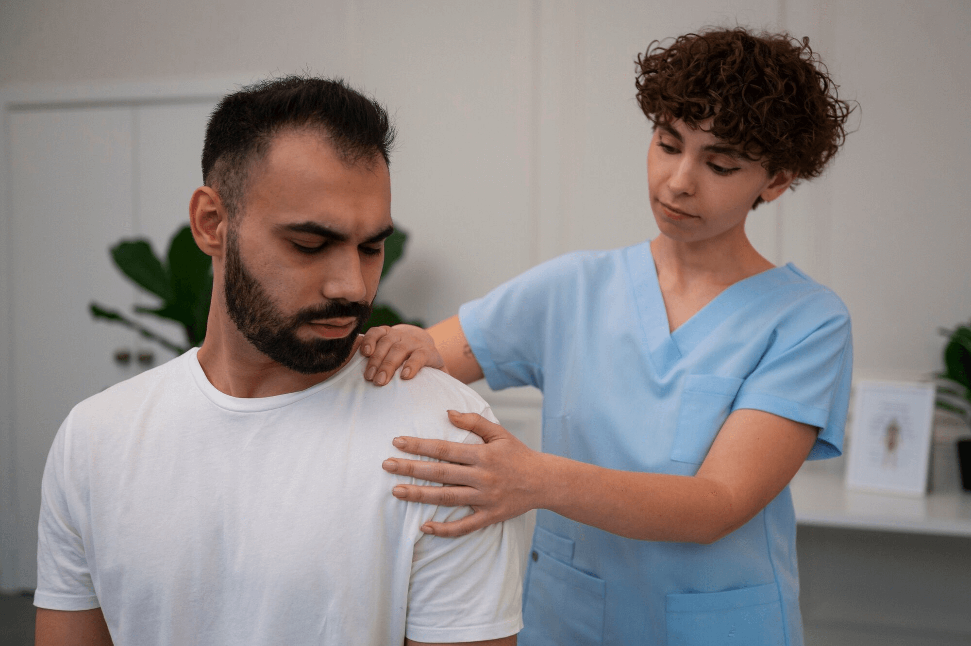 A healthcare professional in blue scrubs examines a seated man's shoulder, possibly addressing back pain from a recent car accident.