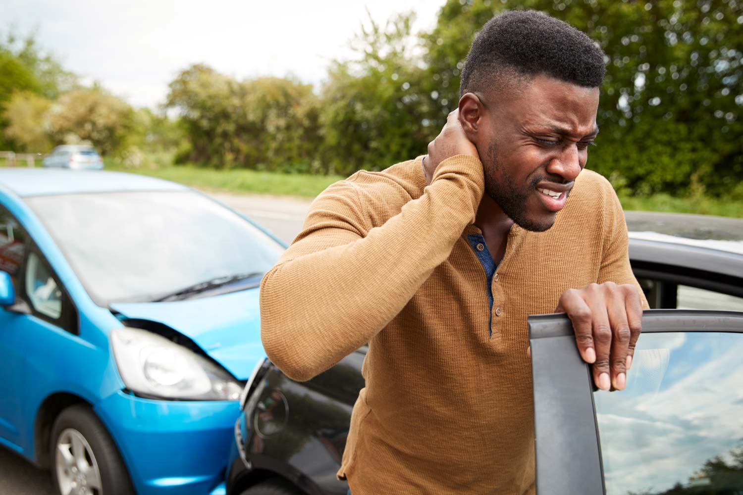 Man holding his neck in pain near his black car after a car accident with a blue car on a road with trees in the background, possibly needing to see a chiropractor for his back pain.