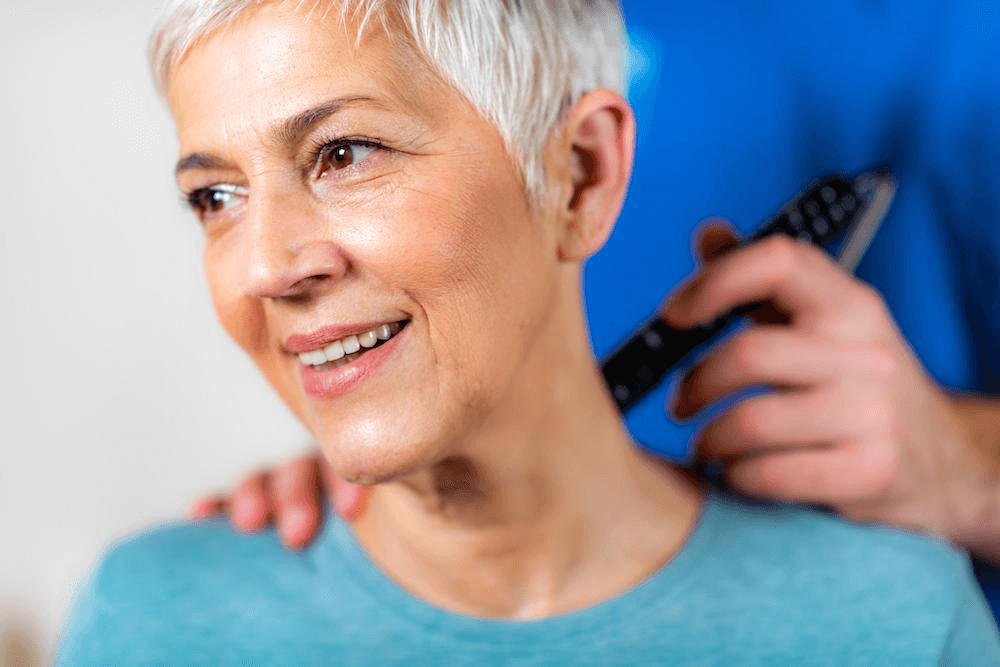 A smiling elderly woman with short gray hair receives a shoulder massage from a chiropractor in a blue shirt.