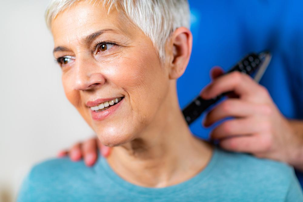 An older woman with short gray hair smiles while receiving a therapeutic treatment from a chiropractor in blue scrubs, holding a handheld device near her neck. Following a recent car accident, the care is helping to alleviate her back pain.